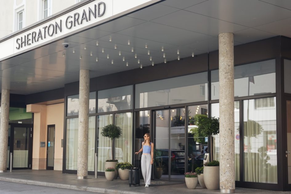 A girl with a carryon luggage standing at the entrance of the hotel with the sign "Sheraton Grand" in Salzburg 
