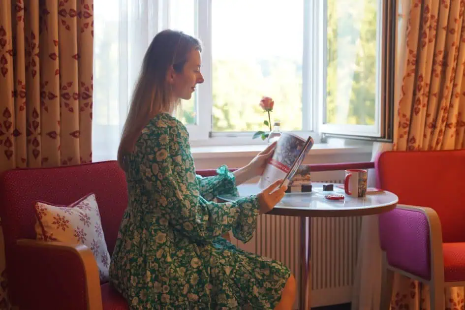 A lady in a green dress sitting on a hotel sofa by the window and reading the magazine 