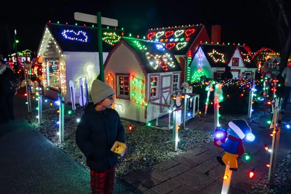 A little boy holding a popcorn standing in front of a lighten tiny houses at the Koziars