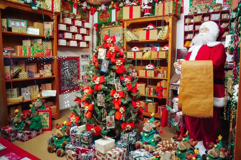 One of the window displays at Koziars featuring Santa with a long gift list in his hand. there is a christmas tree decorated in red bow with gifts underneath and around the shelves