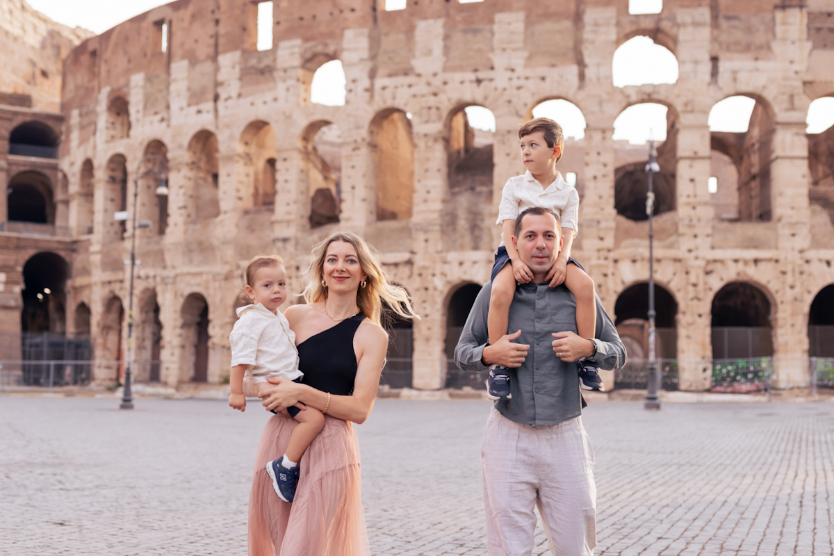 mom and dad carrying two little boys walking towards camera smiling with the background of Colosseum